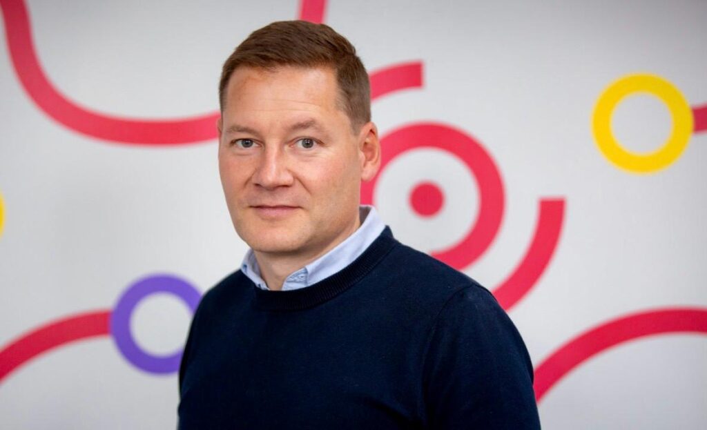 Andy Brown, Director of Propel Tech stands facing the camera, wearing a navy blue crew-neck jumper over a light blue collared shirt, in front of a white wall decorated with colourful ring and swirl graphics in red, yellow, and purple.