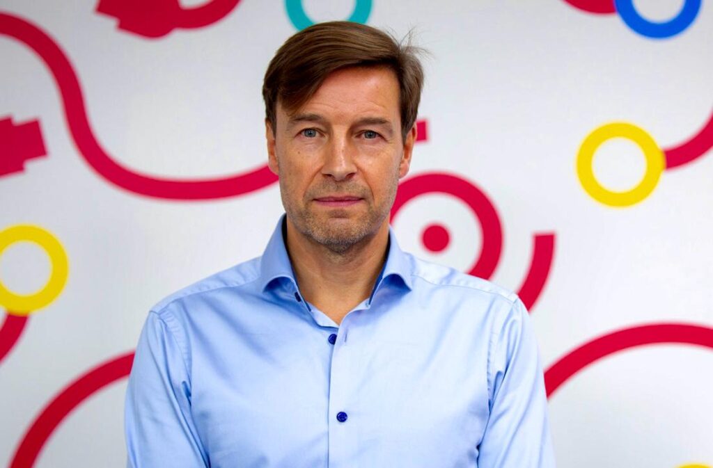 David Ritchie, Director or Propel Tech stands facing the camera, wearing a light blue button-up shirt, in front of a white wall decorated with colourful ring and swirl graphics in red, yellow, and blue