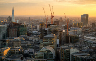 A city skyline at golden hour, with several orange construction cranes rising above a mix of modern glass office buildings and older stone architecture, hazy hills visible in the distance.
