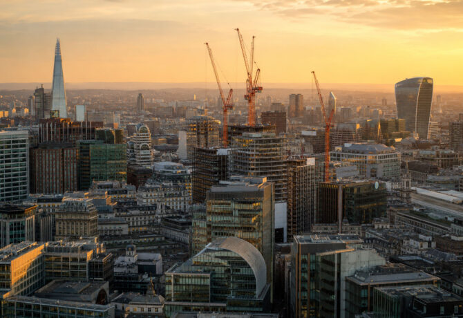 A city skyline at golden hour, with several orange construction cranes rising above a mix of modern glass office buildings and older stone architecture, hazy hills visible in the distance.