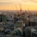 A city skyline at golden hour, with several orange construction cranes rising above a mix of modern glass office buildings and older stone architecture, hazy hills visible in the distance.